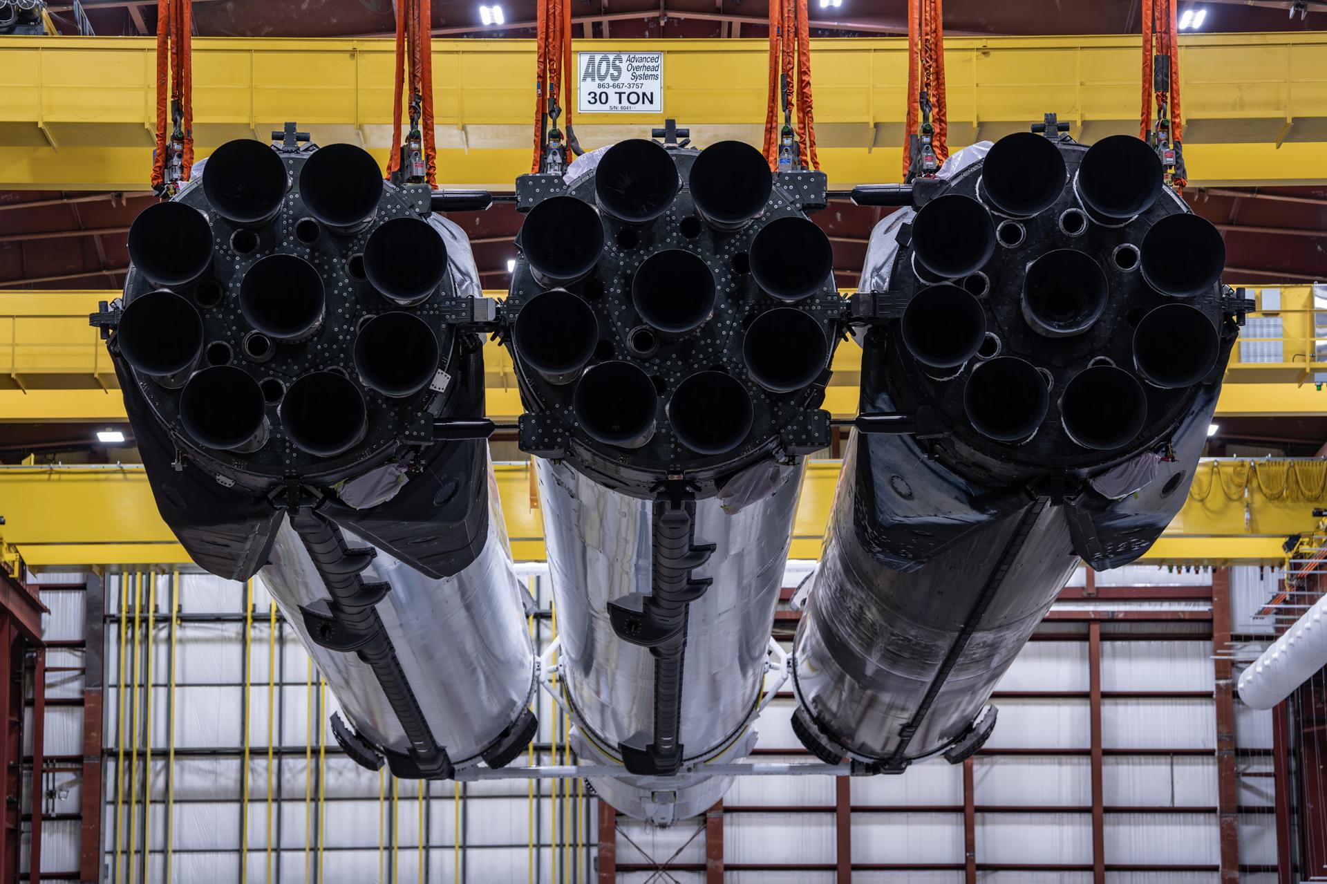 A SpaceX Falcon Heavy rocket is lifted by a yellow crane inside an industrial facility. The 27 black rocket engines stick out of the bottom or three white booster housings, some covered with soot.