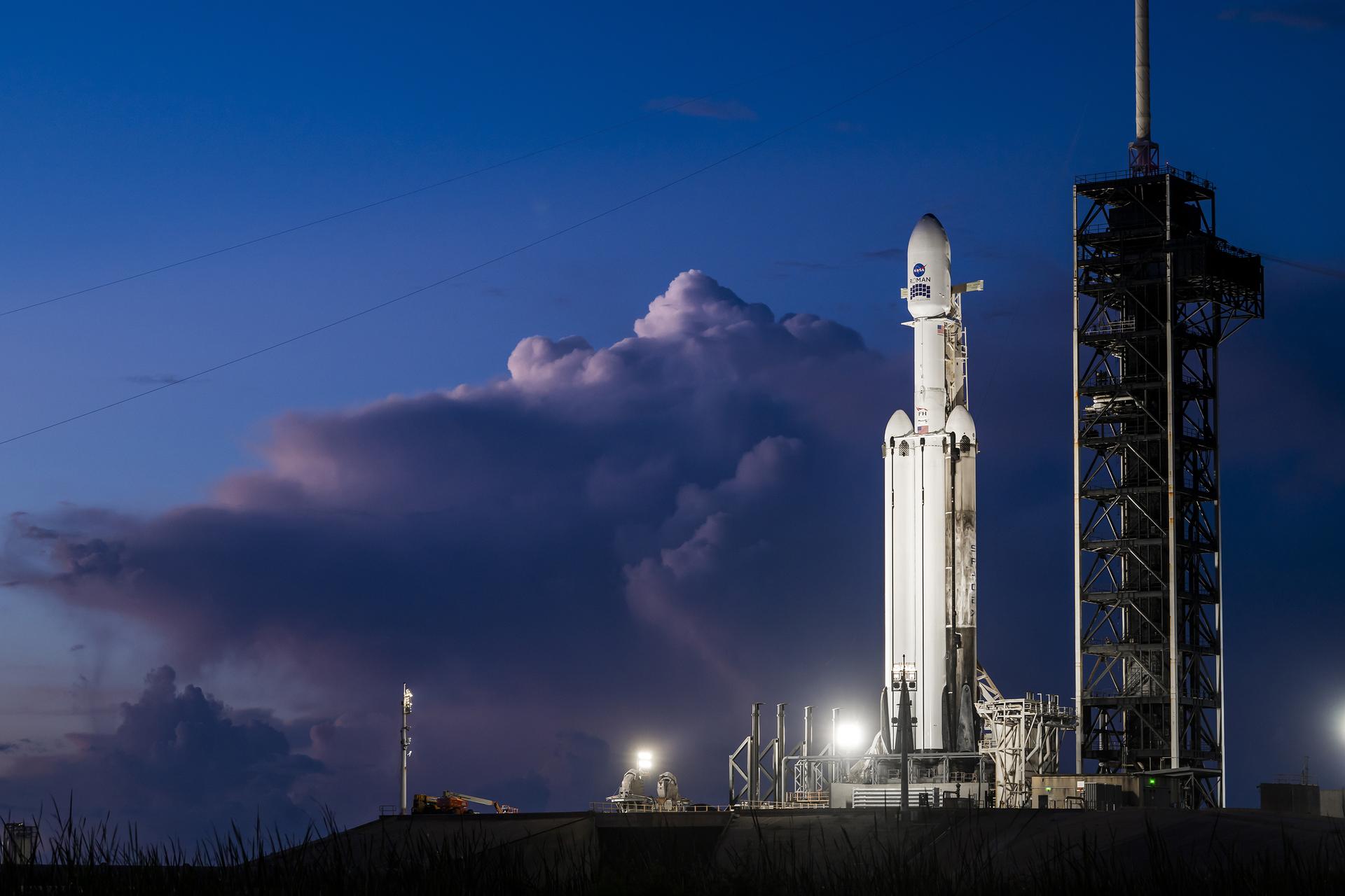 A white SpaceX Falcon Heavy rocket stands vertical on the black launch pad with a predawn sky in the background.