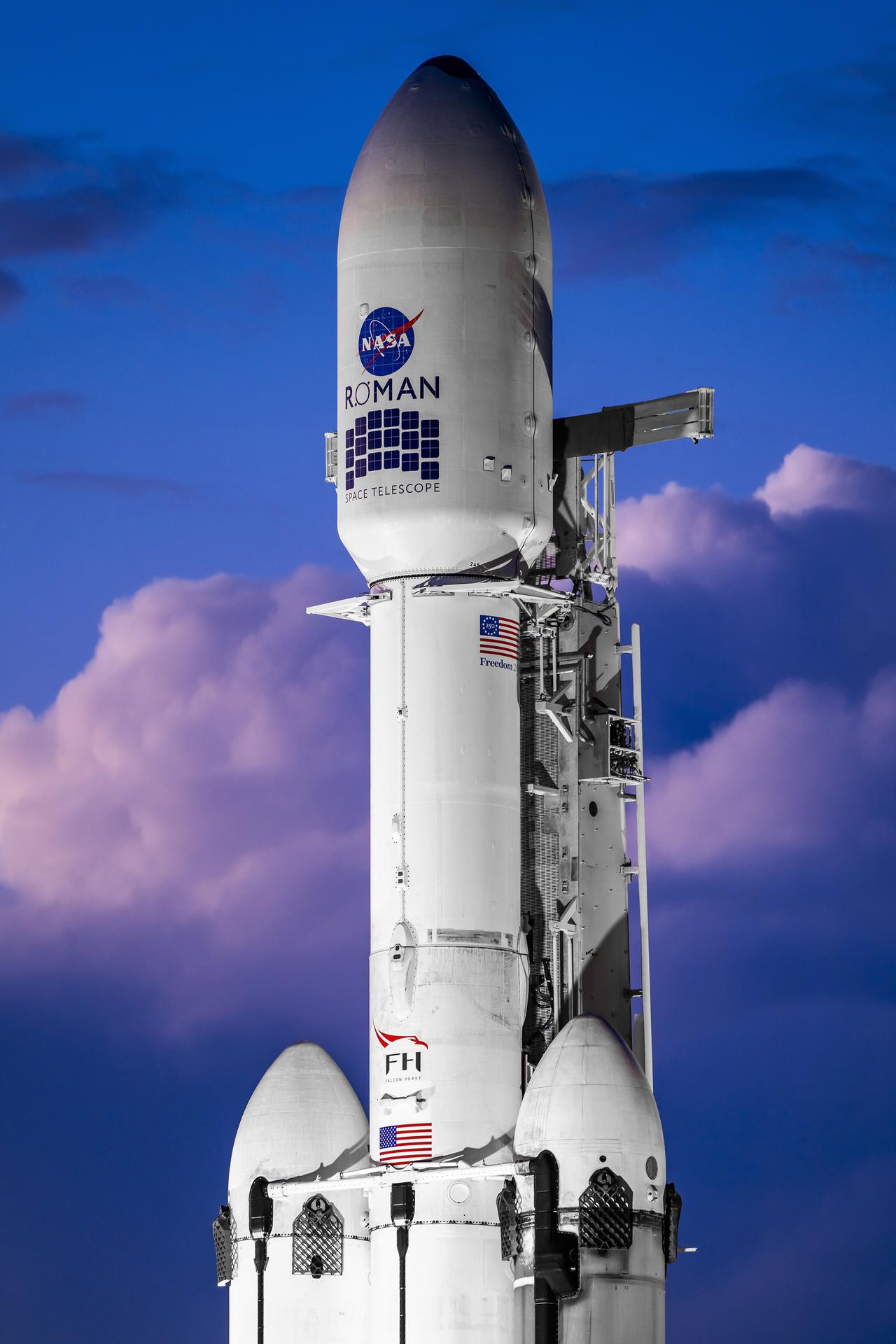 A white SpaceX Falcon Heavy rocket stands vertical on the black launch pad with a predawn sky in the background.