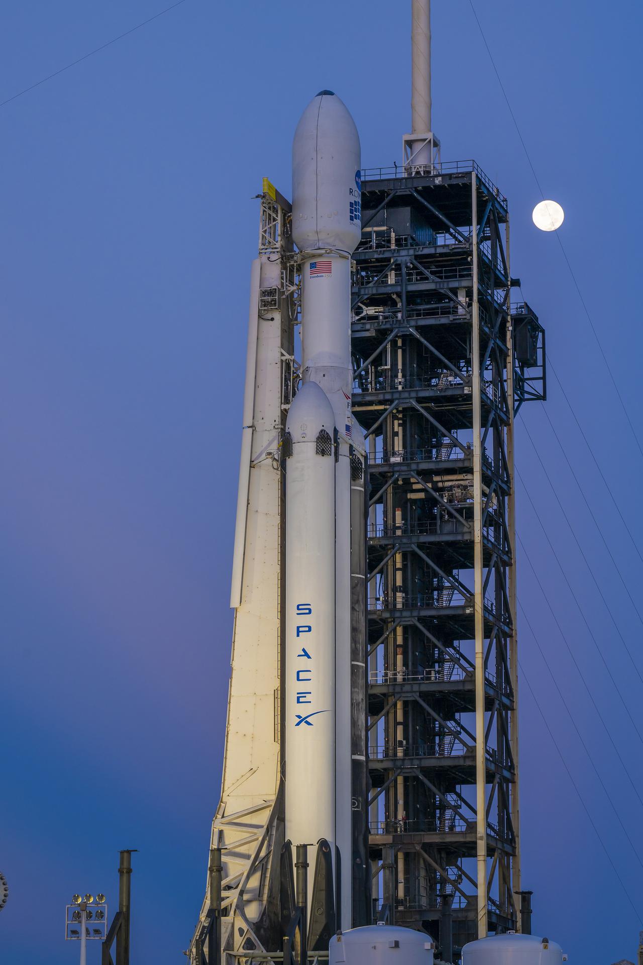 A white SpaceX Falcon Heavy rocket stands vertical on the black launch pad with a predawn sky in the background.
