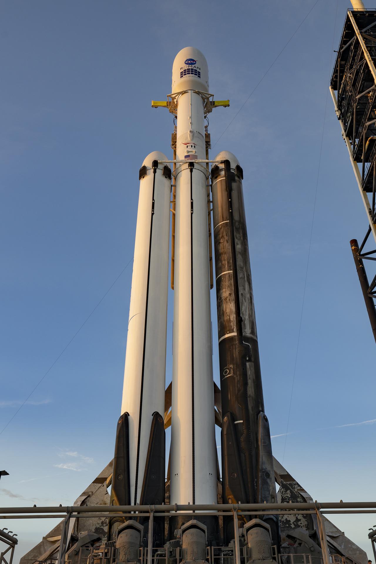 A white SpaceX Falcon Heavy rocket stands vertical on the black launch pad with a predawn sky in the background.