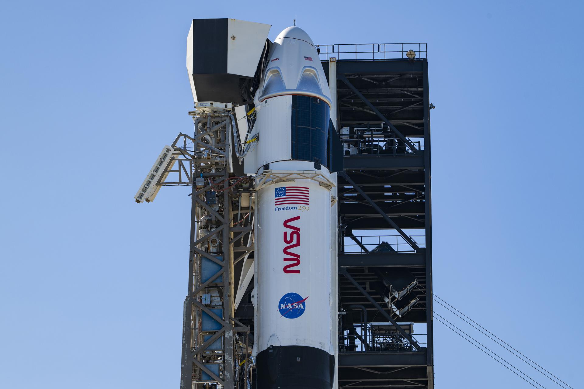 A close-up view of a SpaceX Crew Dragon spacecraft mounted atop a Falcon 9 rocket on the launch pad. The capsule and trunk are connected to ground support hardware, with NASA and mission insignia visible on the rocket’s exterior against a bright blue sky.
