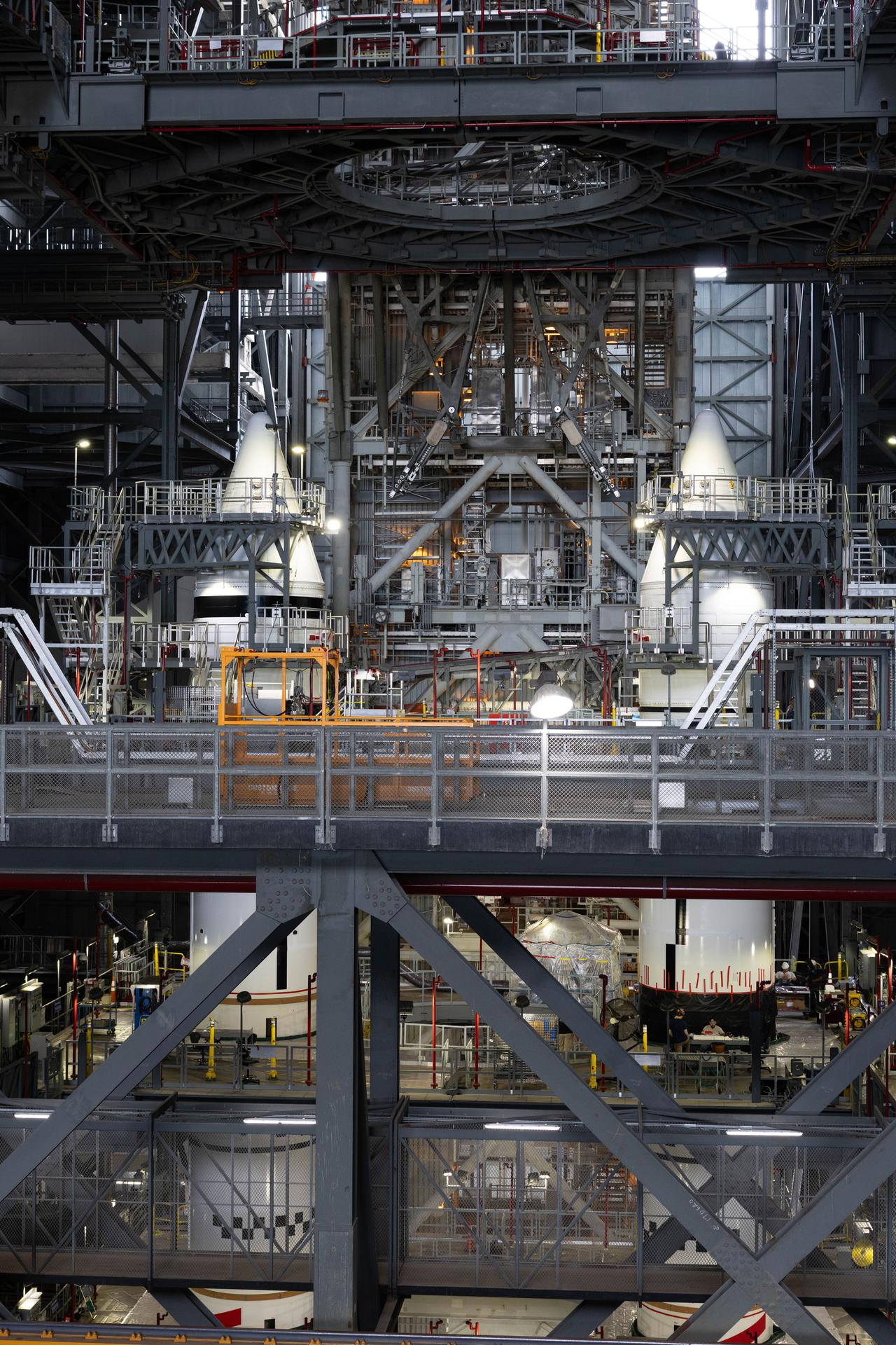 Inside a large industrial facility, two white rocket booster segments stand upright on the ground level, surrounded by scaffolding, platforms, and bright work lights. Two pointy nose cones pieces of hardware atop the booster segments.