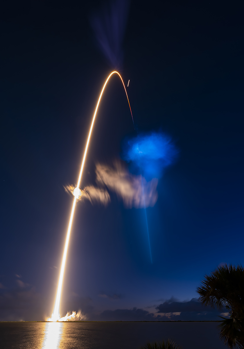 Long-exposure photograph showing a rocket's illuminated flight path and expanding exhaust plume against a dark blue night sky.