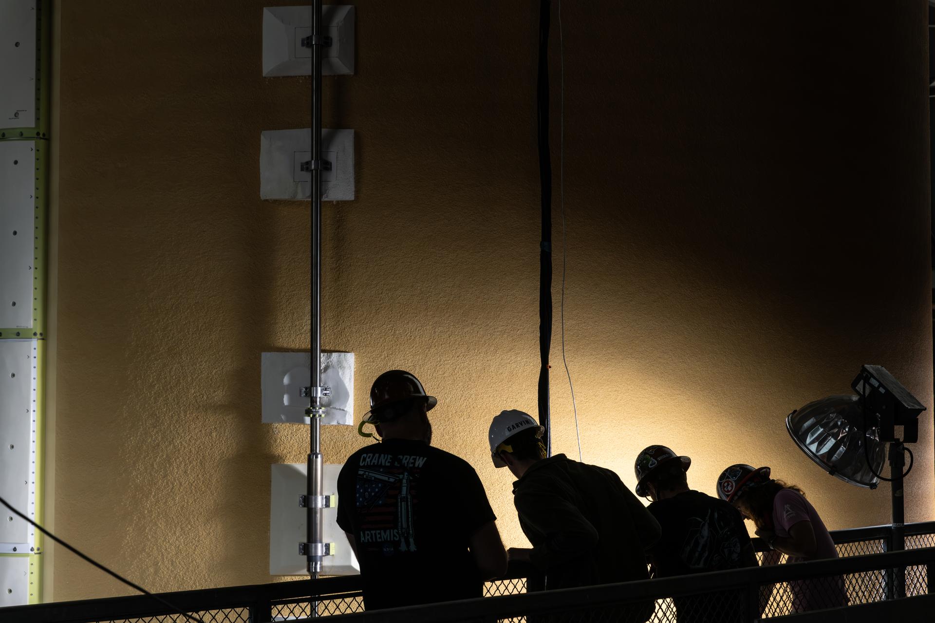 Four people in hard hats are silhouetted against the side of the core stage of the Space Launch System rocket as it is raised in the transfer aisle of the Vehicle Assembly Building. A spotlight on the group’s right shines a bright spot on a portion of the orange stage which is very close and fills the image.