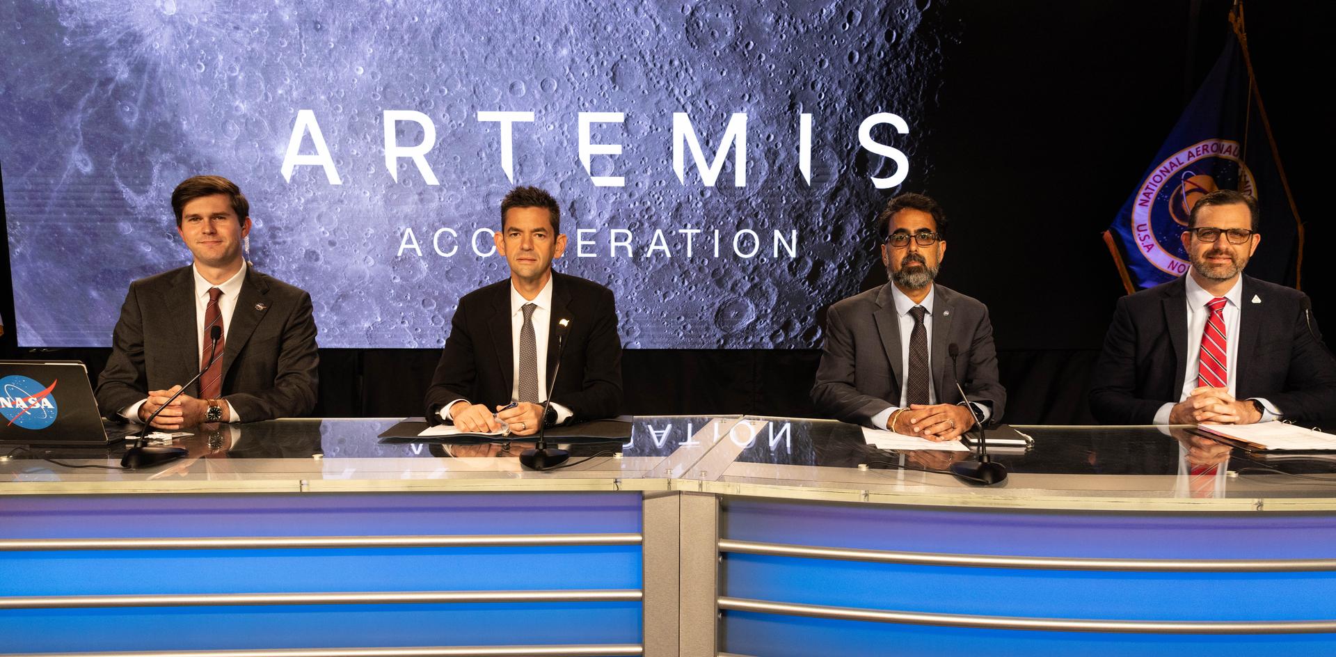 Four people sit behind a curved desk with microphones in a studio setting. A large screen behind them displays the words “ARTEMIS ACCELERATION” over an image of the Moon’s surface. A NASA flag stands at the right side of the stage. The desk is illuminated with blue lighting, and papers and a laptop are visible on the tabletop.