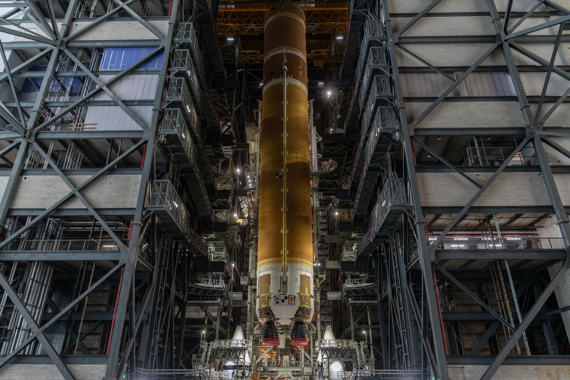 The 212-foot core stage of the Space Launch System rocket is vertical in High Bay 3 of the Vehicle Assembly Building. The nose cones of the two side boosters are visible as the crane places the core stage between them where they are already set on the Mobile Launcher.