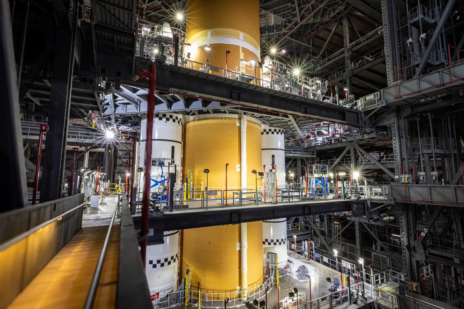 The central portion of the Space Launch System rocket stack is seen from a platform across the central aisle in the Vehicle Assembly Building. The nose cones of the solid rocket booster flank the orange core stage and are surrounded by platforms and staircases.