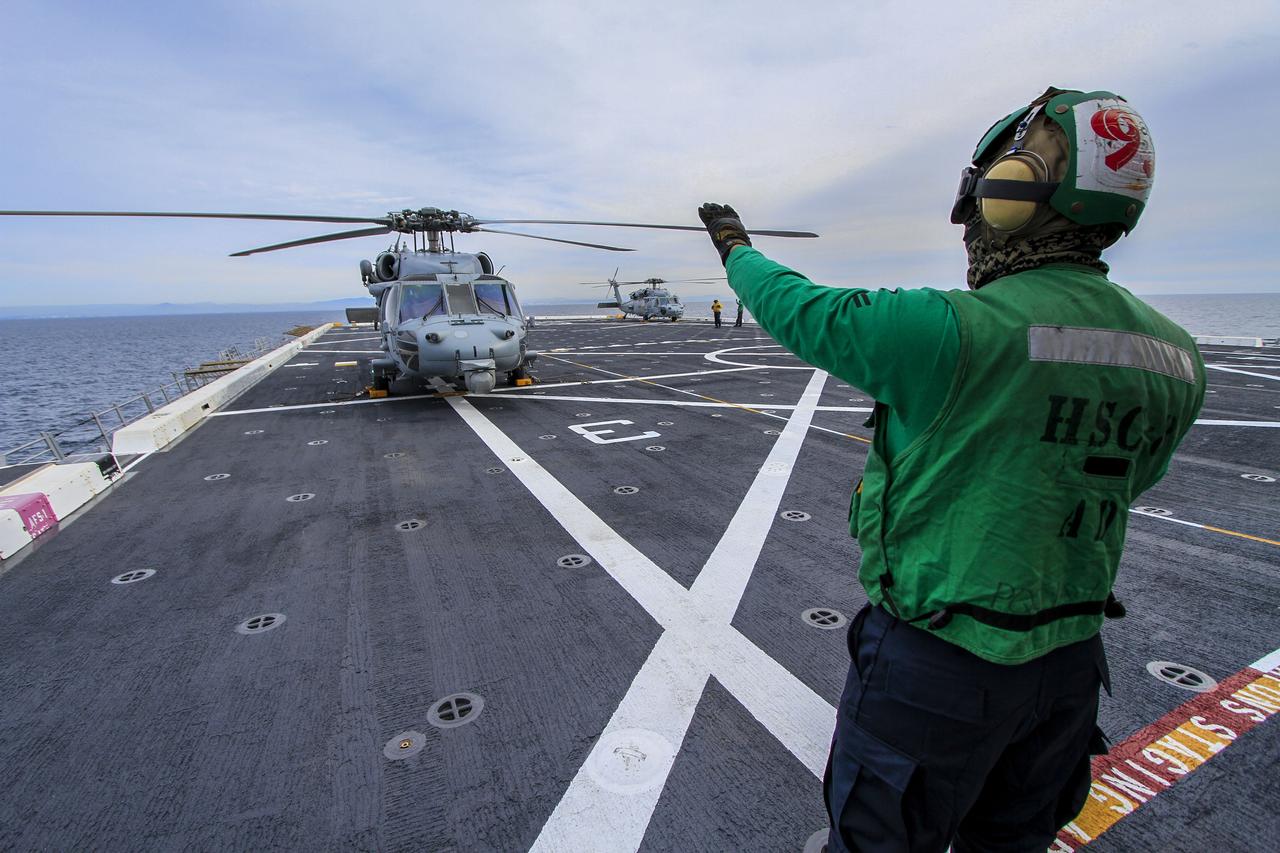 A member of the Helicopter Sea Combat Squadron 8 signals to the pilot in an H60-S Seahawk helicopter on the deck of the USS Anchorage as the ship departs Naval Base San Diego in California for the open waters of the Pacific Ocean. NASA and the U.S. Navy are making preparations ahead of Orion's flight test for recovery of the crew module, forward bay cover and parachutes on its return from space and splashdown in the Pacific Ocean. The Ground Systems Development and Operations Program is leading the recovery efforts. 