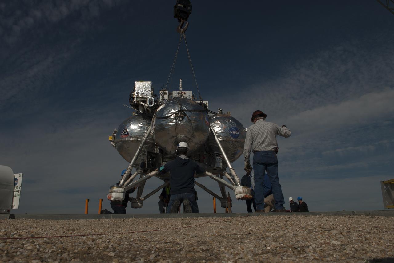 Engineers and technicians prepare NASA's Project Morpheus prototype lander for free flight test number 15 on a launch pad at the north end of the Shuttle Landing Facility at NASA's Kennedy Space Center in Florida. Morpheus is being lowered by crane onto the launch pad. The lander will take off from the ground over a flame trench and use its autonomous landing and hazard avoidance technology, or ALHAT sensors, to survey the hazard field to determine safe landing sites. Project Morpheus tests NASA’s ALHAT and an engine that runs on liquid oxygen and methane, which are green propellants. These new capabilities could be used in future efforts to deliver cargo to planetary surfaces. Project Morpheus is being managed under the Advanced Exploration Systems, or AES, Division in NASA’s Human Exploration and Operations Mission Directorate.