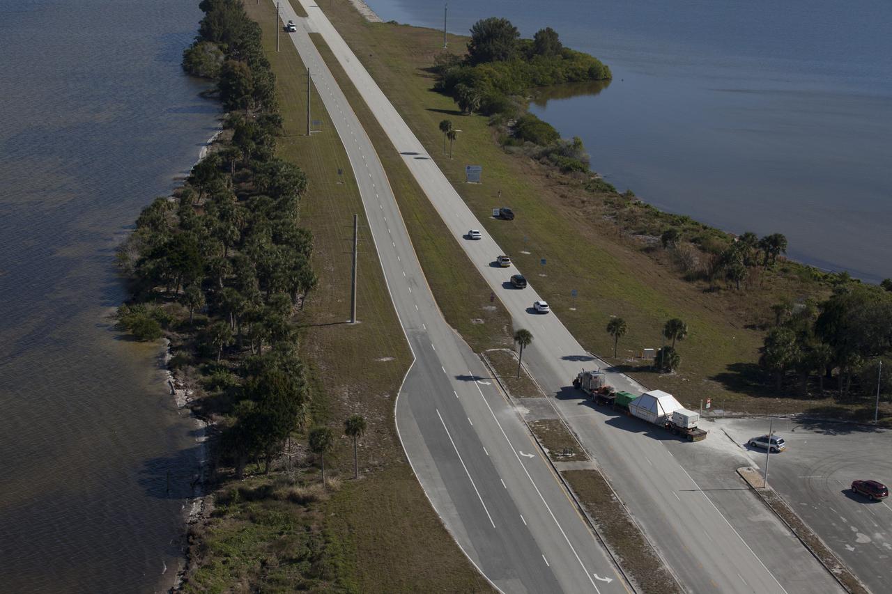 An aerial view near NASA's Kennedy Space Center Visitor Complex reveals the Orion crew module, enclosed in its crew module transportation fixture and secured on a flatbed truck that is proceeding onto the NASA Causeway that leads to the entrance gate to Kennedy Space Center in Florida. Orion made the 2,700 mile overland trip from Naval Base San Diego in California. The spacecraft was recovered from the Pacific Ocean after completing a two-orbit, four-and-a-half hour mission Dec. 5 to test systems critical to crew safety, including the launch abort system, the heat shield and the parachute system. The Ground Systems Development and Operations Program led the recovery, offload and transportation efforts. 