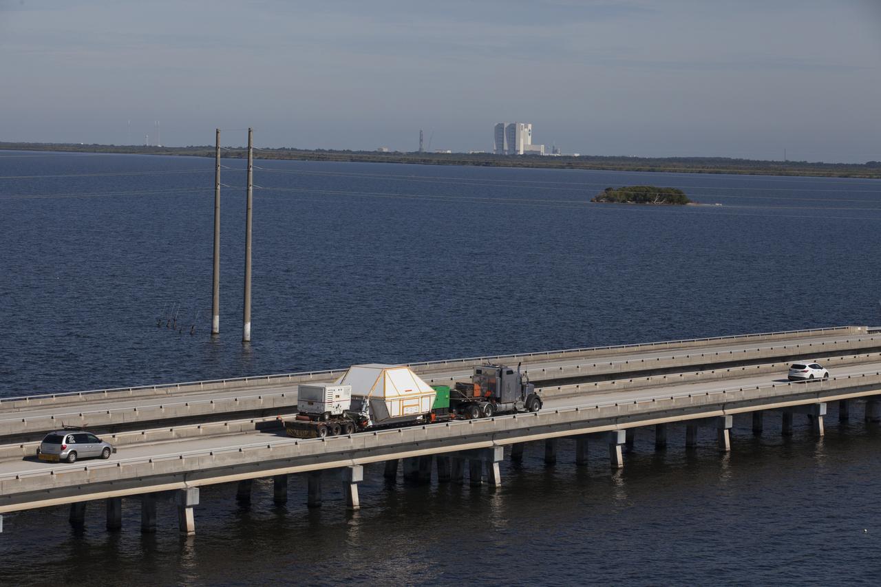 An aerial view near NASA's Kennedy Space Center Visitor Complex reveals the Orion crew module, enclosed in its crew module transportation fixture and secured on a flatbed truck that is proceeding along the NASA Causeway to the entrance gate to Kennedy Space Center in Florida. Across the inland waterway is the iconic Vehicle Assembly Building. Orion made the 2,700 mile overland trip from Naval Base San Diego in California. The spacecraft was recovered from the Pacific Ocean after completing a two-orbit, four-and-a-half hour mission Dec. 5 to test systems critical to crew safety, including the launch abort system, the heat shield and the parachute system. The Ground Systems Development and Operations Program led the recovery, offload and transportation efforts. 