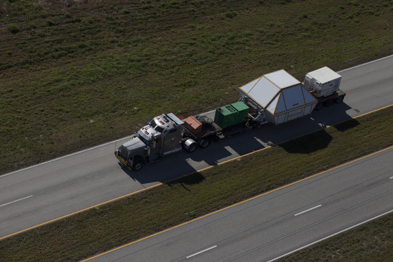 An aerial view near NASA's Kennedy Space Center Visitor Complex reveals the Orion crew module, enclosed in its crew module transportation fixture and secured on a flatbed truck that is proceeding along the NASA Causeway to the entrance gate to Kennedy Space Center in Florida. Orion made the 2,700 mile overland trip from Naval Base San Diego in California. The spacecraft was recovered from the Pacific Ocean after completing a two-orbit, four-and-a-half hour mission Dec. 5 to test systems critical to crew safety, including the launch abort system, the heat shield and the parachute system. The Ground Systems Development and Operations Program led the recovery, offload and transportation efforts.