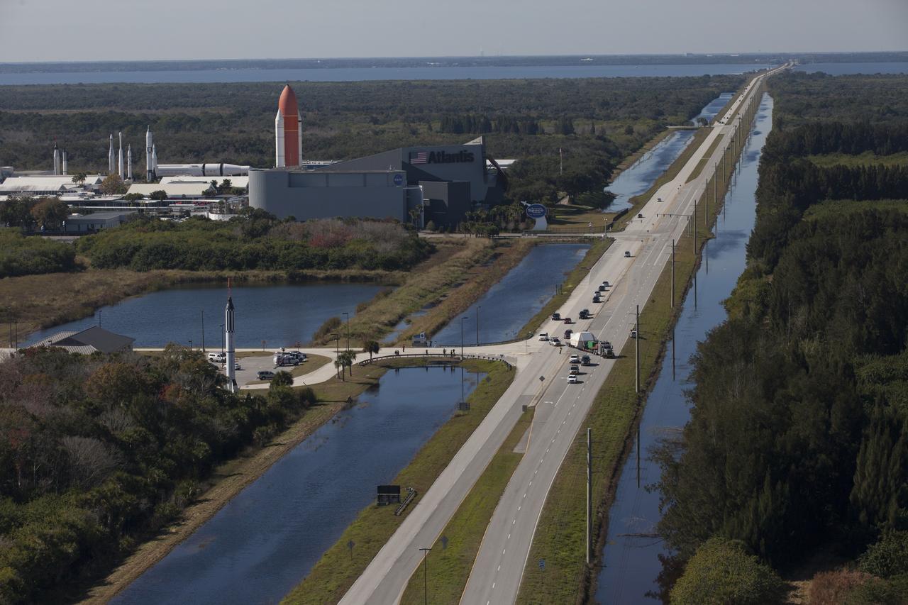In this aerial view, NASA's Orion crew module, enclosed in its crew module transportation fixture and secured on a flatbed truck is passing the Kennedy Space Center Visitor Complex on its way to the entrance gate to Kennedy Space Center in Florida. In view is the Space Shuttle Atlantis facility. Orion made the 2,700 mile overland trip from Naval Base San Diego in California. Orion was recovered from the Pacific Ocean after completing a two-orbit, four-and-a-half hour mission Dec. 5 to test systems critical to crew safety, including the launch abort system, the heat shield and the parachute system. The Ground Systems Development and Operations Program led the recovery, offload and transportation efforts. 