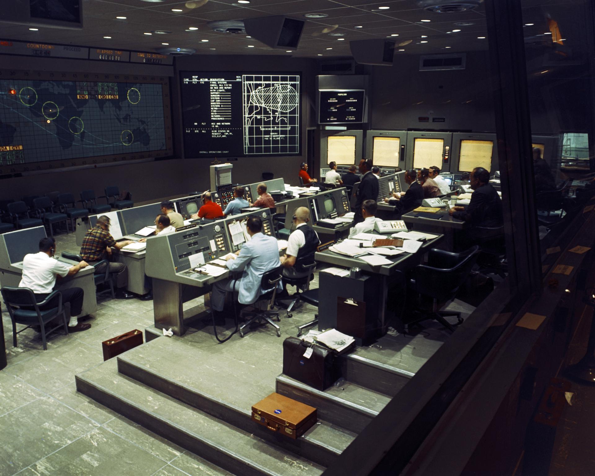 Interior view of the Mission Control Center at Cape Canaveral with flight controllers seated at consoles.