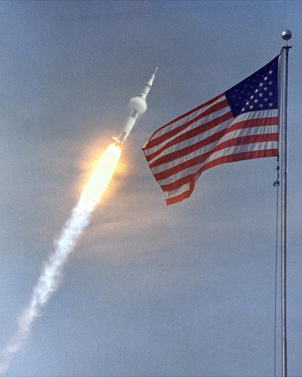 CAPE CANAVERAL, Fla. -- The American Flag heralds the flight of Apollo 11, man's first lunar landing mission. This double exposure was made with a 1,000 mm lens. The photograph was taken from Cape Kennedy, adjacent to Kennedy Space Center, where Apollo 11 lifted off from pad 39A at 9:32 a.m. EDT. This image was imposed upon the image of hte flag, filmed a day earlier. In the photo, the rocket at an alititude of about 5,000 feet. A band of super-cold propellants seems to circle the rocket near its center. The effect is caused by the difference in temperature between the propellants and the atmosphere.    Photo credit: NASA