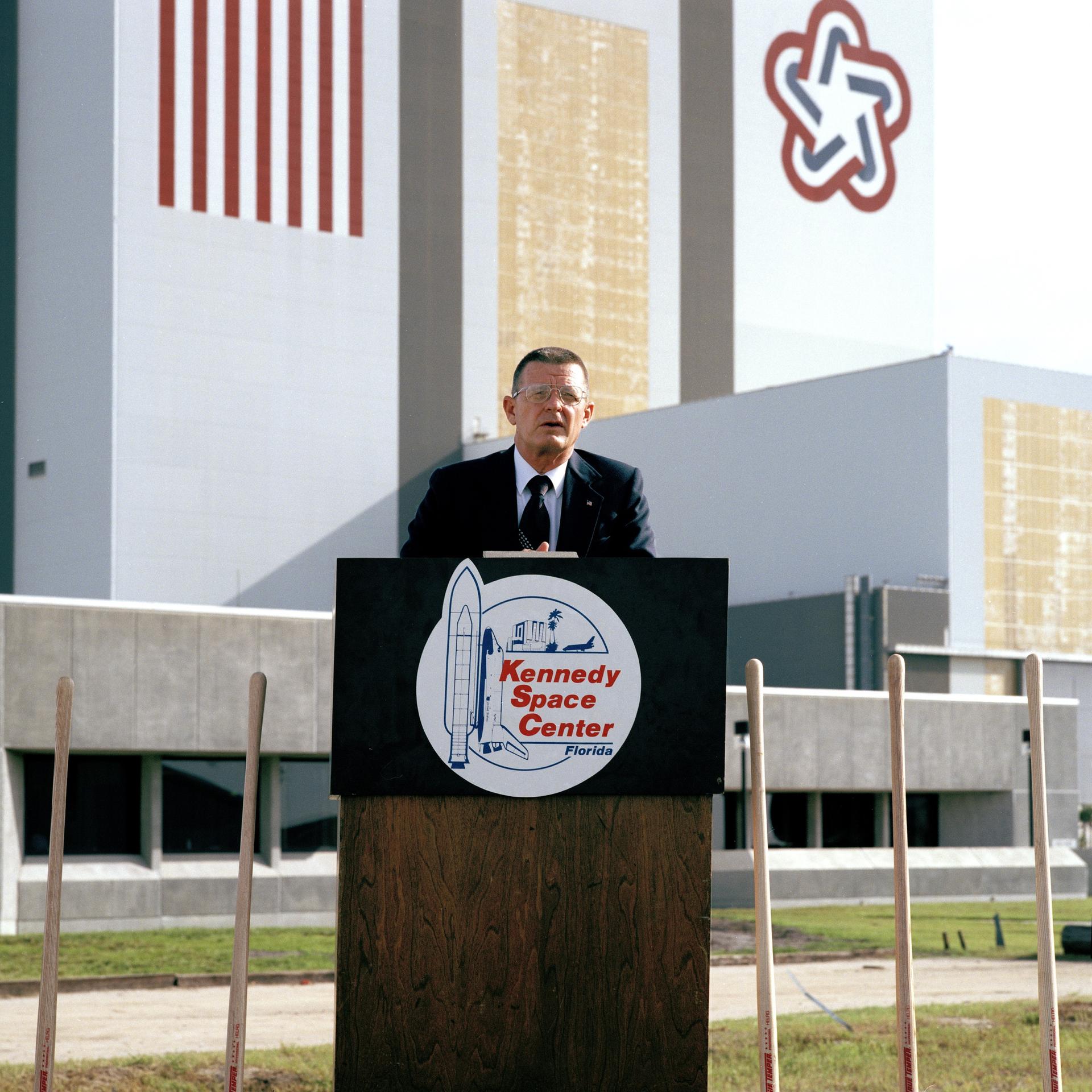 Center Director Forrest McCartney speaks at a podium in front of the Vehicle Assembly Building.