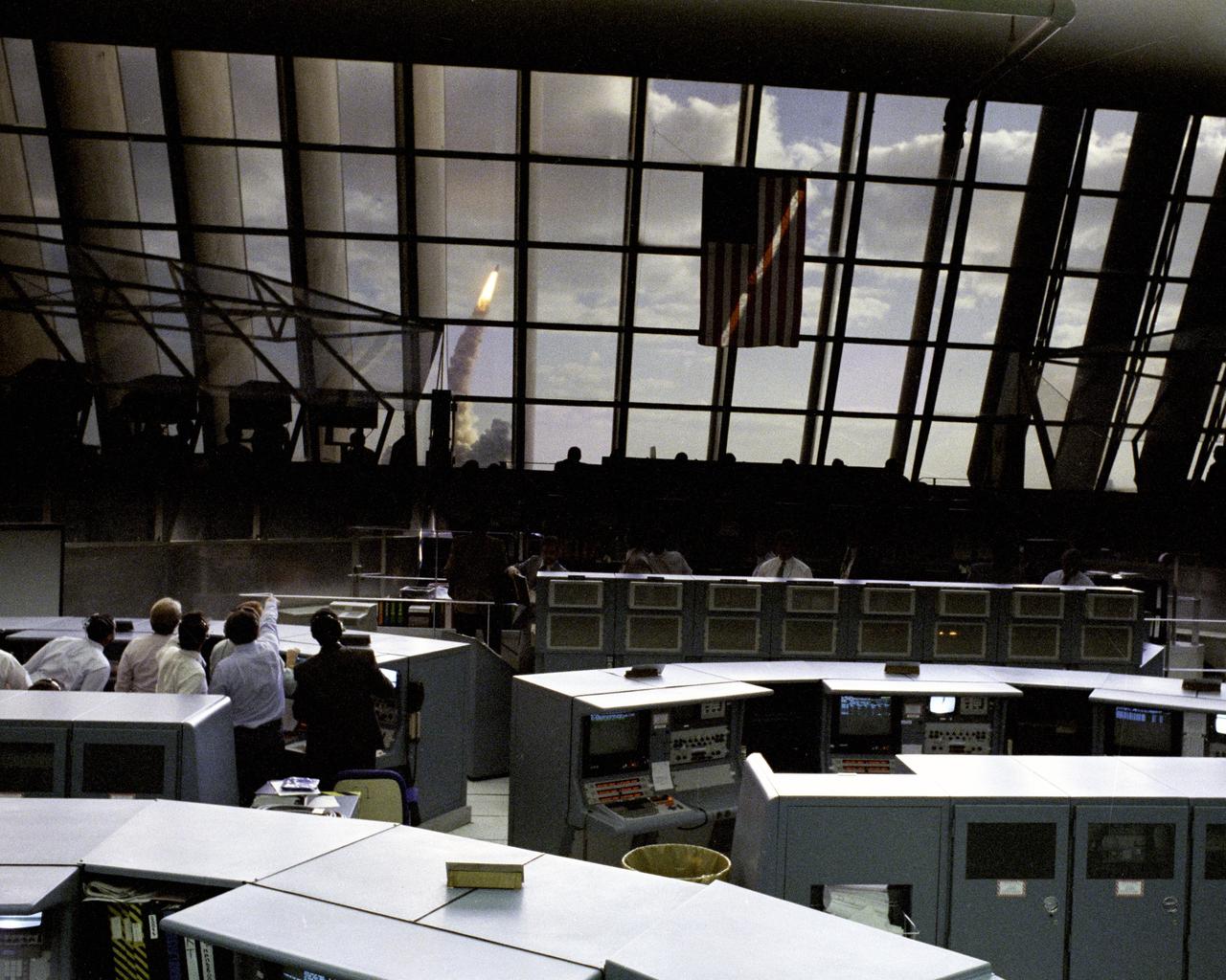 Through the large window panes of Firing Room 1, KSC launch team members reap the rewards of their work with a glimpse of the space shuttle Discovery soaring into the sky. Discovery was launched for the tenth time at 8:34 a.m. EDT on April 24 beginning the five-day STS-31 mission to deploy the Hubble Space Telescope. A ray of morning sunlight highlights the red and white stripes of Old Glory hanging high in the Firing Room. Launch team members overcame a last minute challenge in the STS-31 countdown when software detected a main propulsion system valve was out of position. The situation was quickly corrected and verified by the team from consoles in the Firing Room and the countdown was returned in a matter of minutes. Photo credit: NASA