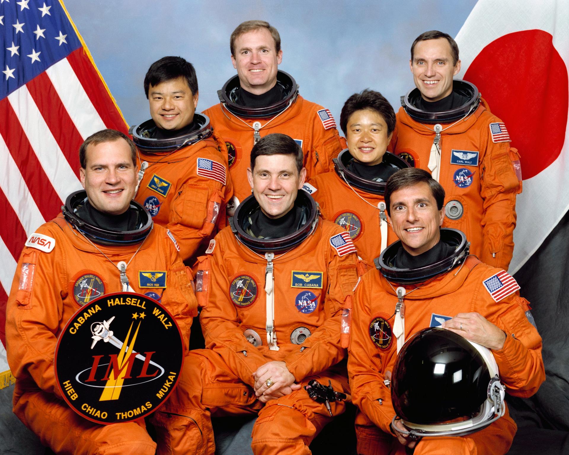 Seven men in orange spacesuits pose for crew photo in front of American and Japanese flag.