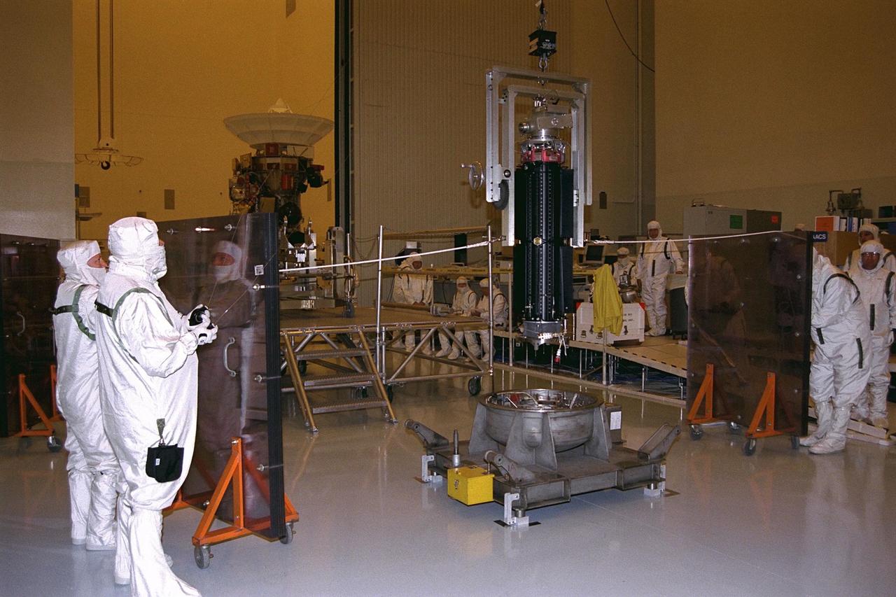 Supported on a lift fixture, this radioisotope  thermoelectric generator (RTG), at center, is hoisted from its storage base using the  airlock crane in the Payload Hazardous Servicing Facility (PHSF). Jet Propulsion  Laboratory (JPL) workers are preparing to install the RTG onto the Cassini spacecraft,  in background at left, for mechanical and electrical verification testing. The three RTGs  on Cassini will provide electrical power to the spacecraft on its 6.7-year trip to the  Saturnian system and during its four-year mission at Saturn. RTGs use heat from the  natural decay of plutonium to generate electric power. The generators enable spacecraft  to operate at great distances from the Sun where solar power systems are not feasible.  The Cassini mission is targeted for an Oct. 6 launch aboard a Titan IVB/Centaur  expendable launch vehicle. Cassini is built and managed by JPL