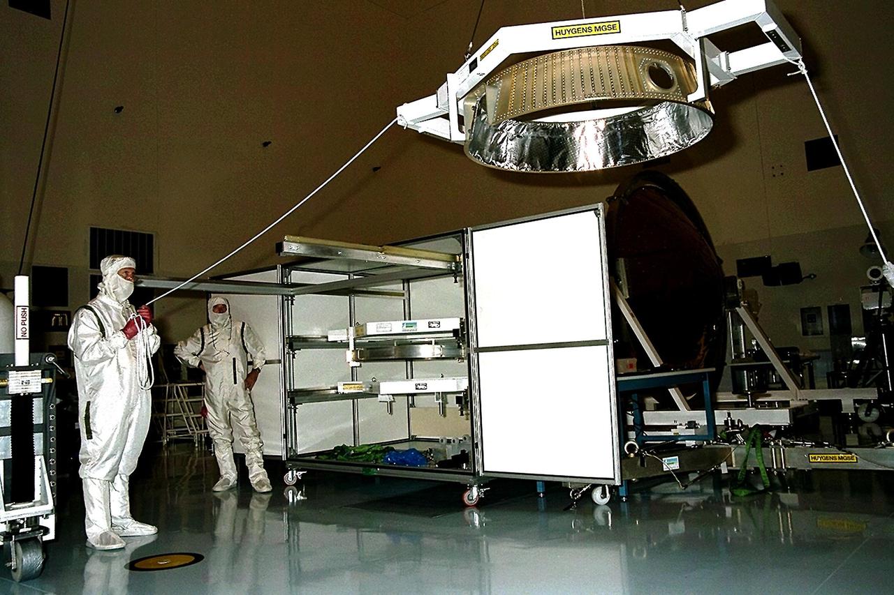 Dornier Satelliten Systeme (DSS) workers lift part of the Huygens probe aft cover assembly in the Payload Hazardous Servicing Facility (PHSF) at KSC. The spacecraft was returned to the PHSF after damage to thermal insulation was discovered inside Huygens from an abnormally high flow of conditioned air. Internal inspection, insulation repair and a cleaning of the probe were required. Mission managers are targeting a mid-October launch date after the Cassini spacecraft, aboard which Huygens will be launched, returns to the pad and is once again placed atop its Titan IVB expendable launch vehicle at Launch Pad 40 at Cape Canaveral Air Station