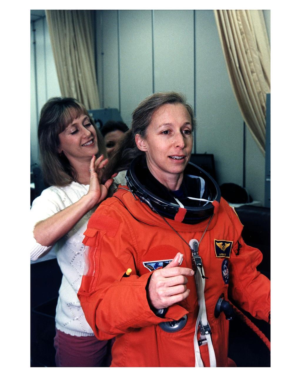 STS-81 Mission Specialist Marsha S. Ivins gets a helping hand from a suit technician as she prepares to don the helmet of her launch/entry suit in the suitup room of the Operations and Checkout (O&C) Building. She is the veteran of three Shuttle flights and became an astronaut in 1984. Among other responsibilities, Ivins will perform photo and video surveys of the Russian Mir space station and operate the Kidsat experiment camera on the orbiter’s aft flight deck. She and five crew members will shortly depart the O&C and head for Launch Pad 39B, where the Space Shuttle Atlantis will lift off during a 7-minute window that opens at 4:27 a.m. EST, January 12