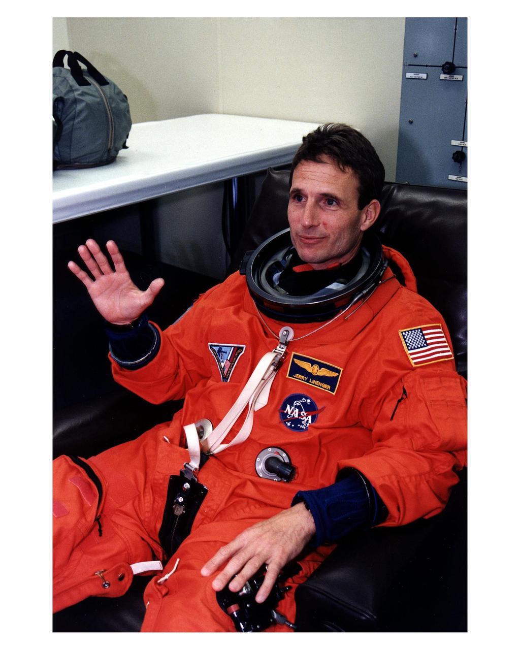 STS-81 Mission Specialist Jerry Linenger waves to the camera in his launch/entry suit and helmet in the suitup room of the Operations and Checkout (O&C) Building. He is on his second Shuttle flight and has been an astronaut since 1992. Linenger will become a member of the Mir 22 crew and replace astronaut John Blaha on the Russian space station for a four-month stay after the Space Shuttle orbiter Atlantis docks with the orbital habitat on flight day 3. A medical doctor and an exercise buff, Linenger will conduct physiological experiments during his stay on Mir. He and five crew members will shortly depart the O&C and head for Launch Pad 39B, where the Space Shuttle Atlantis will lift off during a 7-minute window that opens at 4:27 a.m. EST, January 12