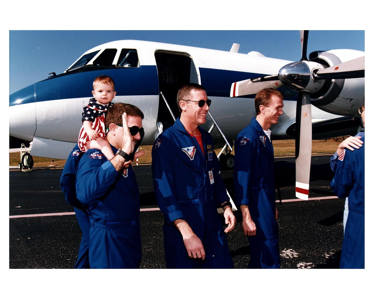 Members of the STS-81 crew prepare to depart for Johnson Space Center Jan. 23 from the Skid Strip at Cape Canaveral Air Station. The crew arrived at KSC aboard the Space Shuttle Atlantis Jan. 22 to conclude the fifth Shuttle-Mir docking mission and return U. S. astronaut John E. Blaha to Earth after four months in space as member of the Mir 22 crew. On the left is Mission Specialist John M. Grunsfeld, with his daughter, Sarah, on his back. Also pictured are Mission Commander Michael A. Baker (center); Pilot Brent W. Jett (second from right); and Blaha