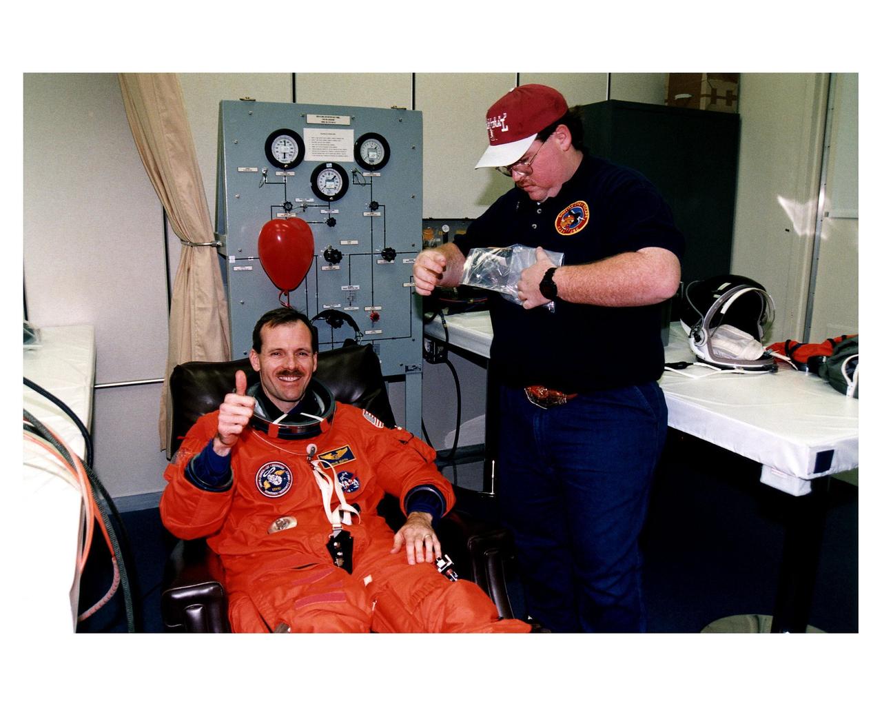 STS-82 Mission Specialist Steven L. Smith gives a "thumbs up" while donning his launch and entry suit in the Operations and Checkout Building. A suit technician stands ready to assist with final adjustments. This is Smith’s second space flight. He and the six other crew members will depart shortly for Launch Pad 39A, where the Space Shuttle Discovery awaits liftoff on a 10-day mission to service the orbiting Hubble Space Telescope (HST). This will be the second HST servicing mission. Four back-to-back spacewalks are planned