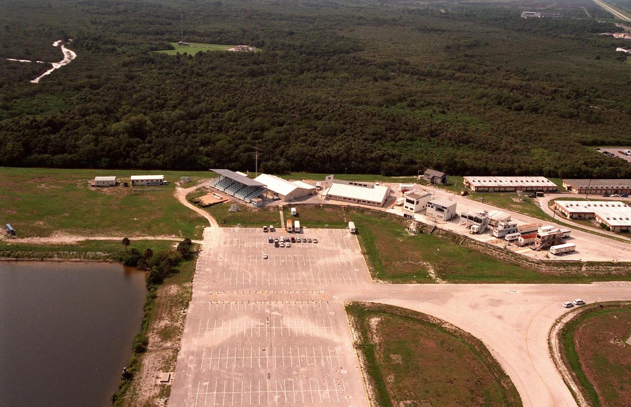In this aerial view, The News Center sits beyond a large parking lot, on a hill at the northeastern end of the Launch Complex 39 Area , next to the turn basin (at left). From left, the grandstand faces the launch pads several miles away on the Atlantic seashore; behind it, the television studio is the site of media conferences; next, the large white-roofed building is the hub of information and activity for press representatives. Lined up on the right of the Press Site are various buildings and trailers, home to major news networks. The parking lot can accommodate the hundreds of media personnel who attend Space Shuttle launches