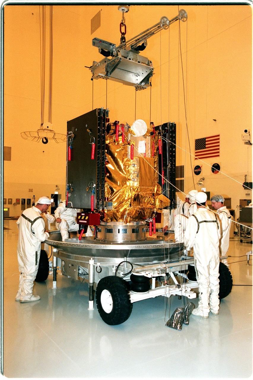KENNEDY SPACE CENTER, FLA. -- Workers in the Payload Hazardous Servicing Facility lower Deep Space 1 onto its transporter, for movement to the Defense Satellite Communications System Processing Facility (DPF), Cape Canaveral Air Station, where it will undergo testing. At either side of the spacecraft are its solar wings, folded for launch. When fully extended, the wings measure 38.6 feet from tip to tip. The first flight in NASA's New Millennium Program, Deep Space 1 is designed to validate 12 new technologies for scientific space missions of the next century. Onboard experiments include a solar-powered ion propulsion engine and software that tracks celestial bodies so the spacecraft can make its own navigation decisions without the intervention of ground controllers. The ion propulsion engine is the first non-chemical propulsion to be used as the primary means of propelling a spacecraft. Deep Space 1 will complete most of its mission objectives within the first two months, but may also do a flyby of a near-Earth asteroid, 1992 KD, in July 1999. Deep Space 1 will be launched aboard a Boeing Delta 7326 rocket from Launch Pad 17A, Cape Canaveral Air Station, in October. Delta II rockets are medium capacity expendable launch vehicles derived from the Delta family of rockets built and launched since 1960. Since then there have been more than 245 Delta launches