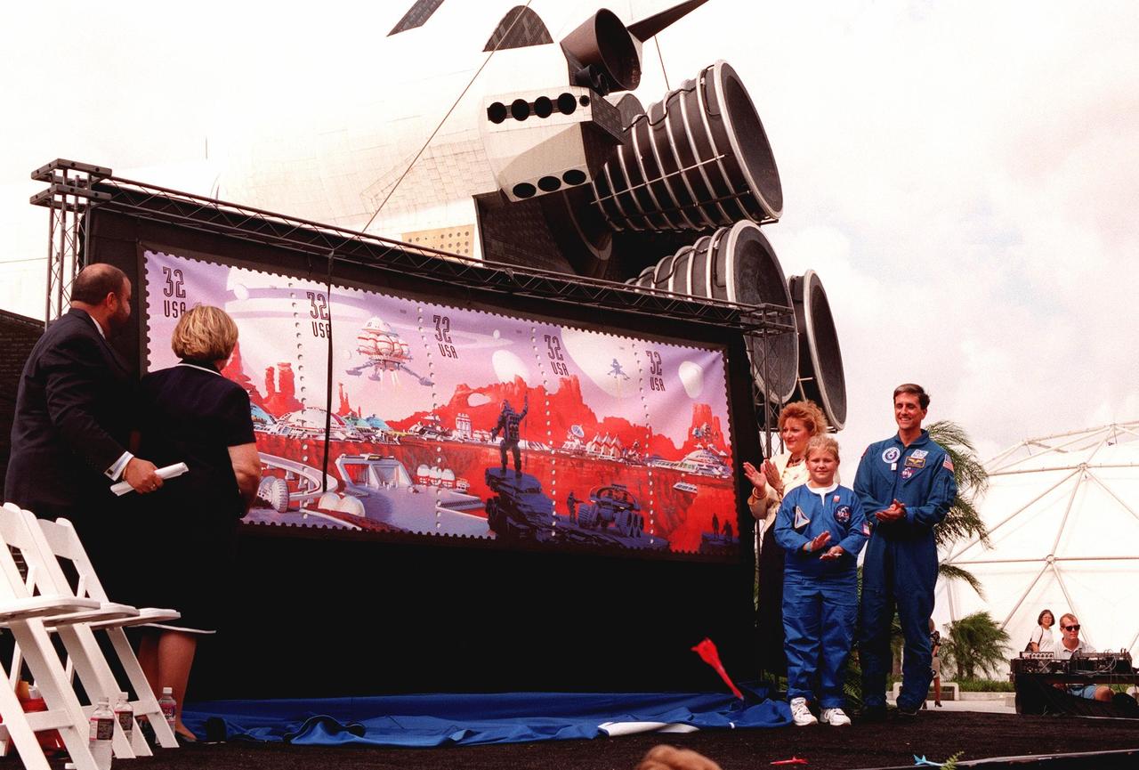 Applauding the unveiling of the U.S. Postal Service's newest series of stamps, Space Discovery, are are (left to right) Barry Ziehl, U.S. Postal Service; JoAnn Morgan, KSC associate director of advanced development and shuttle upgrades; Karla Corcoran, Postal Service inspector general; Kristene A. Graves, a student from Lewis Carroll Elementary School; and Dr. Donald Thomas, astronaut and veteran of four Shuttle missions. During the ceremony, Kristene read her essay "My Stamp Adventure" that she had written for an area-wide contest for the event. The unveiling took place at the KSC Visitor Complex and coincided with NASA's 40th anniversary on this date. Behind the large display can be seen the mockup of an orbiter. The stamps were designed by renowned aerospace artist Attila Heija. The strip of five individual stamps together make up a futuristic scene complete with space vehicles, a futuristic space city, and space explorers. The stamps are available nationwide beginning Oct. 1