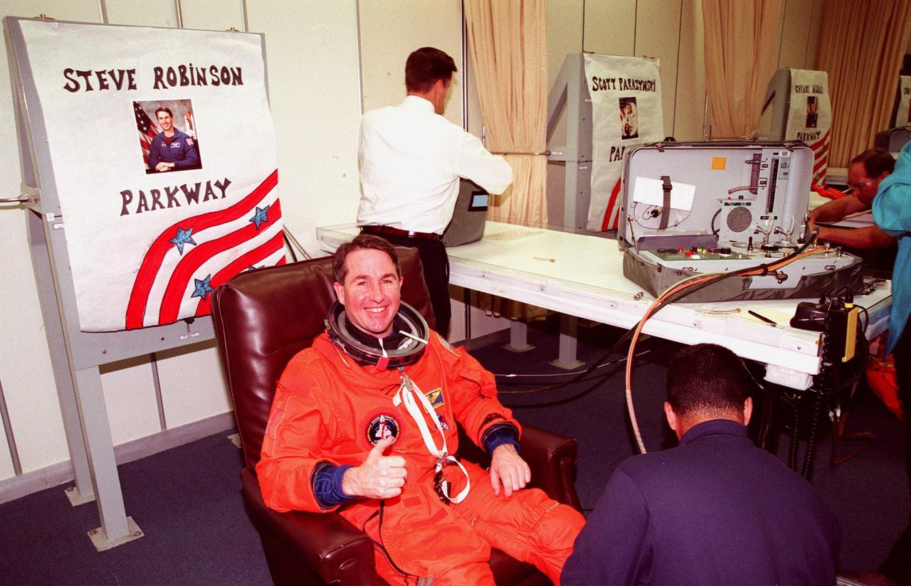 STS-95 Mission Specialist Stephen K. Robinson gives a thumbs up as he dons his flight suit in the Operations and Checkout Building with the help of suit tech George Brittingham (lower right). The final fitting takes place prior to the crew walkout and transport to Launch Pad 39B. Targeted for launch at 2 p.m. EST on Oct. 29, the mission is expected to last 8 days, 21 hours and 49 minutes, and return to KSC at 11:49 a.m. EST on Nov. 7. The STS-95 mission includes research payloads such as the Spartan solar-observing deployable spacecraft, the Hubble Space Telescope Orbital Systems Test Platform, the International Extreme Ultraviolet Hitchhiker, as well as the SPACEHAB single module with experiments on space flight and the aging process