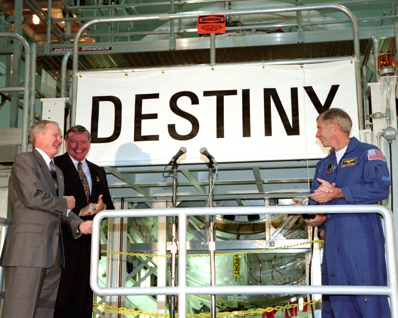 KENNEDY SPACE CENTER, FLA. -  In the Space Station Processing Facility, Center Director Roy Bridges (left), Program Manager of the International Space Station (ISS) Randy Brinkley (second from left) and STS-98 Commander Ken Cockrell (right) applaud the unveiling of the name "Destiny" for the U.S. Laboratory module.  The lab, which is behnd them on a workstand, is scheduled to be launched on STS-98 on Space Shuttle Endeavour in early 2000.  It will become the centerpiece of scientific research on the ISS.  The Shuttle will spend six days docked to the Station while the laboratory is attached and three spacewalks are conducted to compete its assembly.  The laboratory will be launched with five equipment racks aboard, which will provide essential functions for Station systems, including high data-rate communications, and maintain the Station's orientation using control gyroscopes launched earlier.  Additional equipment and research racks will be installed in the laboratory on subsequent Shuttle flights.