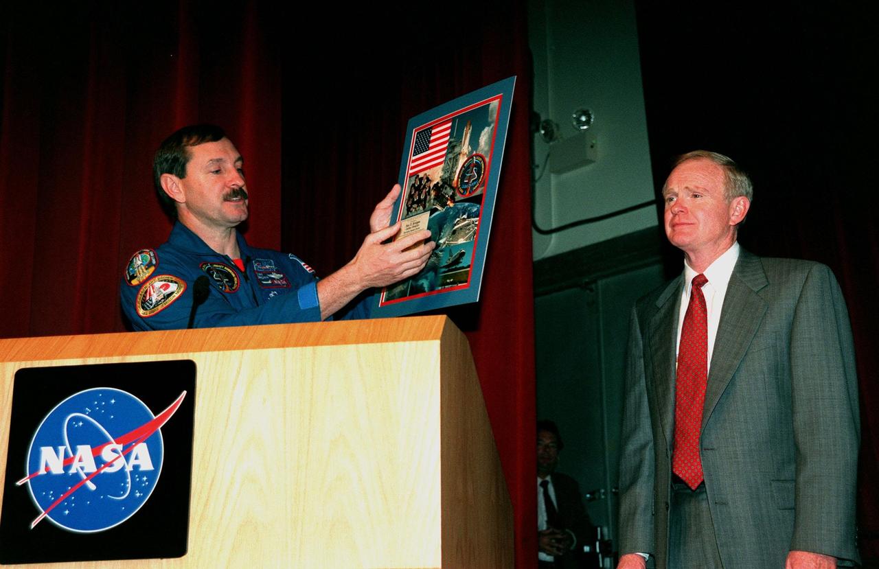 STS-95 Commander Curtis L. Brown Jr. (left) presents a composite photograph of images taken during the STS-95 mission to Kennedy Space Center (KSC) Director Roy Bridges in the Training Auditorium. The auditorium is filled with KSC employees who were invited to hear the crew describe their experiences during their successful mission dedicated to microgravity research and to view a videotape of the highlights of the mission. The other crew members are Pilot Steven W. Lindsey; Mission Specialist and Payload Commander Stephen K. Robinson; Mission Specialists Scott E. Parazynski and Pedro Duque, with the European Space Agency (ESA); and Payload Specialists Chiaki Mukai, with the National Space Development Agency of Japan (NASDA), and John H. Glenn Jr., a senator from Ohio and one of the original seven Project Mercury astronauts. Later in the afternoon, the crew will participate in a parade down State Road A1A in nearby Cocoa Beach, reminiscent of those held after missions during the Mercury Program