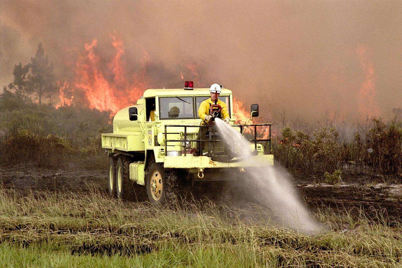 KENNEDY SPACE CENTER, FLA. --  A fire burns in the background as members of the U.S Fish and Wildlife Service operate firefighting equipment soaking the grass and underbrush in an attempt to keep the fire away from Kennedy Parkway and the wooded area on the other side of the road. Lightning touched off three different fires Sunday evening in and around Kennedy Space Center at Tel IV, Ransom Road and Pine Island Road. This area is part of the Merritt Island National Wildlife Refuge operated by the service. The fires were a short distance from operational facilities at the space center and forced the closing of Florida State Route 3. The fires are being contained by firefighters from Kennedy Space Center and the U.S. Fish and Wildlife Service