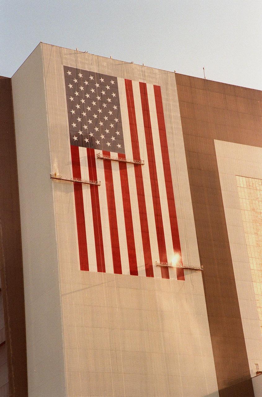 Painters are suspended on platforms from the top of the 525-foot-high Vehicle Assembly Building (VAB) at KSC during repainting of the American flag. The flag spans an area 209 feet by 110 feet and will require 510 gallons of red, white and blue paint. Each stripe of the flag is 9 feet wide and each star is 6 feet in diameter. The platforms are operated by two electric motors and travel 35 feet per minute. Work is being done with rollers, with brushes being used for details. The paint was donated by ICI Devoe of Louisville, Ky. In addition to the flag, the Bicentennial Emblem on the other side of the VAB doors is being replaced by the NASA logo, honoring NASA’s 40th anniversary (in October). The logo covers an area 110 feet by 132 feet. Work is expected to be completed in mid-September