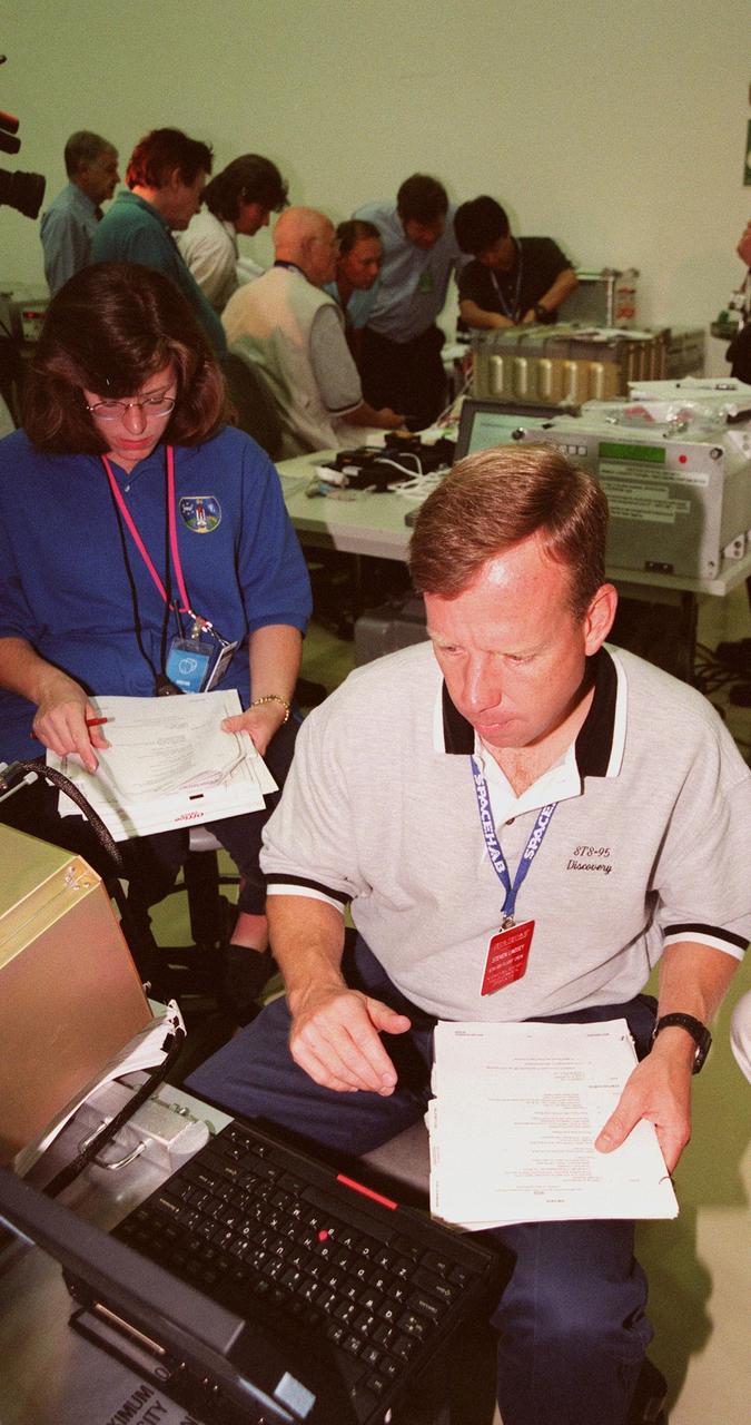 KENNEDY SPACE CENTER, FLA. -- In the SPACEHAB Payload Processing Facility (SPPF), Cape Canaveral, STS-95 Pilot Steven Lindsey (right) and a KSC worker (left) look over documentation, while other crew members (in the background) check out equipment. Lindsey and the rest of the crew have been participating in SPACEHAB familiarization in the SPPF. The mission includes research payloads such as the Spartan solar-observing deployable spacecraft, the Hubble Space Telescope Orbital Systems Test Platform, the International Extreme Ultraviolet Hitchhiker, as well as the SPACEHAB single module with experiments on space flight and the aging process