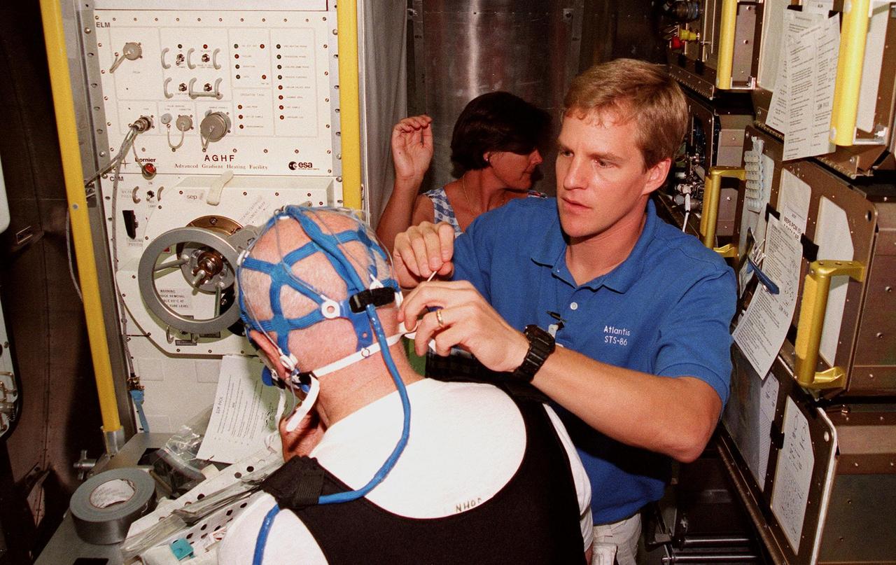 KENNEDY SPACE CENTER, FLA. -- Inside the SPACEHAB training module, STS-95 Mission Specialist Scott Parazynski, M.D. (right), attaches sensors to the mesh cap worn by Payload Specialist John Glenn (back to camera). In the background is Ann Elliott, University of California, San Diego. Glenn will wear the cap on the mission to monitor and record brain waves during sleep. Parazynski and Glenn are participating in SPACEHAB familiarization at the SPACEHAB Payload Processing Facility, Cape Canaveral. The mission, scheduled to launch Oct. 29, includes research payloads such as the Spartan solar-observing deployable spacecraft, the Hubble Space Telescope Orbital Systems Test Platform, the International Extreme Ultraviolet Hitchhiker, as well as the SPACEHAB single module with experiments on space flight and the aging process