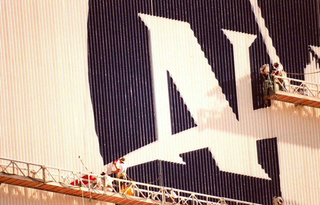 KENNEDY SPACE CENTER, FLA. -- The worker on the lower left applies the red paint to the chevron while the worker on the right fills in the blue field to the NASA logo they are painting on the Vehicle Assembly Building (VAB). When finished, the logo, also known as the "meatball," will measure 110 feet by 132 feet, or about 12,300 square feet. Workers, suspended on platforms from the top of the 525-foot-high VAB, are using rollers and brushes to do the painting. The entire fleet of orbiters is also receiving the addition of the NASA logo on their wings and sidewalls. In addition to the logo, the American flag is being repainted on the other side of the VAB. The flag spans an area 209 feet by 110 feet, or about 23, 437 square feet. Each stripe is 9 feet wide and each star is 6 feet in diameter. The painting honors NASA’s 40th anniversary on Oct. 1 and is expected to be complete in mid-September