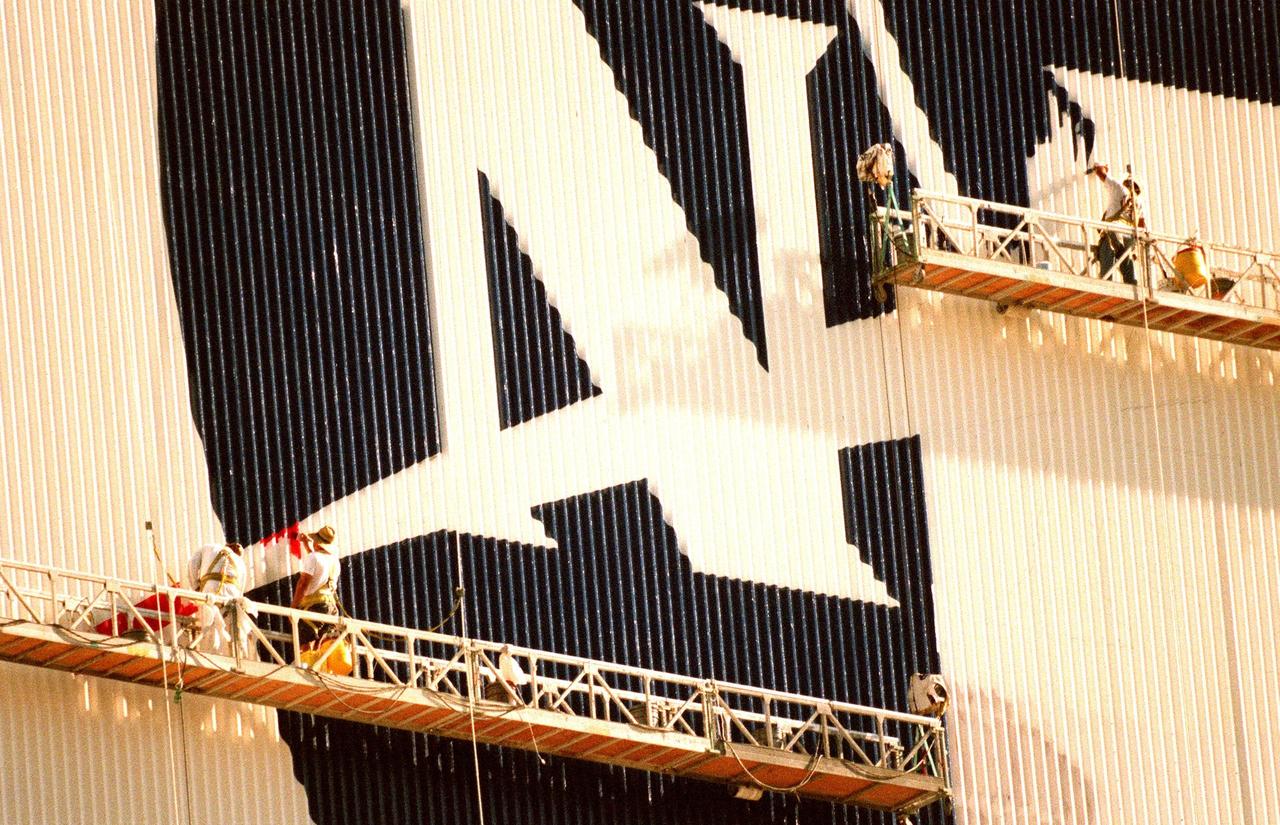 KENNEDY SPACE CENTER, FLA. -- The worker on the lower left applies the red paint to the chevron while the worker on the right fills in the blue field to the NASA logo they are painting on the Vehicle Assembly Building (VAB). When finished, the logo, also known as the "meatball," will measure 110 feet by 132 feet, or about 12,300 square feet. Workers, suspended on platforms from the top of the 525-foot-high VAB, are using rollers and brushes to do the painting. The entire fleet of orbiters is also receiving the addition of the NASA logo on their wings and sidewalls. In addition to the logo, the American flag is being repainted on the other side of the VAB. The flag spans an area 209 feet by 110 feet, or about 23, 437 square feet. Each stripe is 9 feet wide and each star is 6 feet in diameter. The painting honors NASA’s 40th anniversary on Oct. 1 and is expected to be complete in mid-September