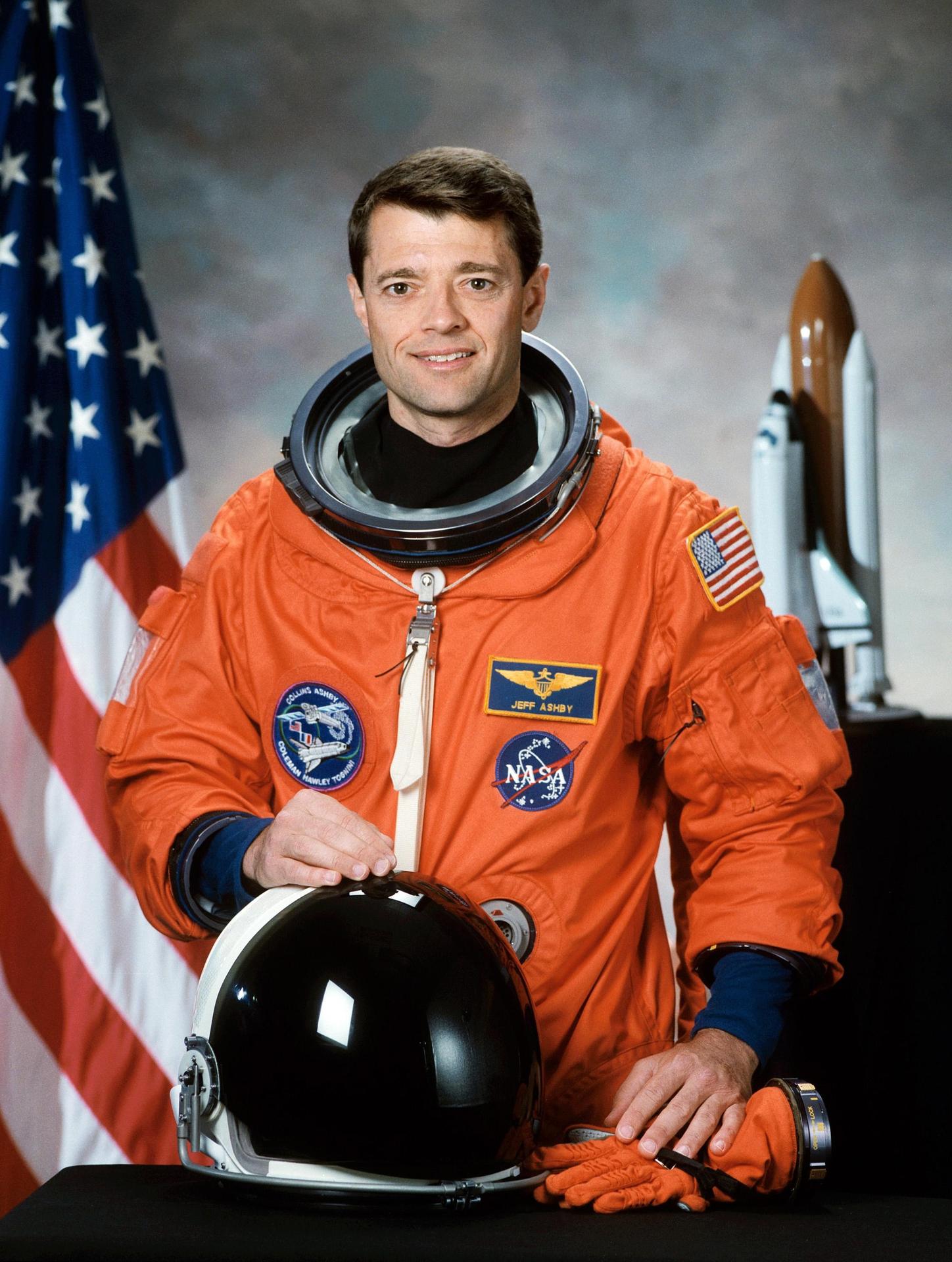 A man sits before the American flag in an astronaut suite, with his helmet off