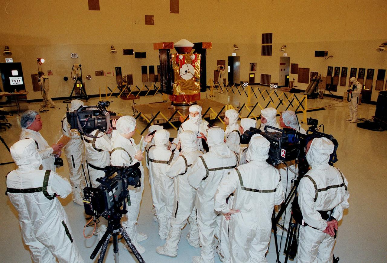In the Payload Hazardous Servicing Facility, media representatives, dressed in protective suits, are updated by Project Manager Richard Grammier (center, top), with the Jet Propulsion Laboratory, about the Stardust spacecraft (in the background). Stardust is targeted for launch on Feb. 6 aboard a Boeing Delta II rocket from Launch Pad 17-A, Cape Canaveral Air Station. The spacecraft is destined for a close encounter with the comet Wild 2 in January 2004. Using a silicon-based substance called aerogel, Stardust will capture comet particles flying off the nucleus of the comet. The spacecraft also will bring back samples of interstellar dust. These materials consist of ancient pre-solar interstellar grains and other remnants left over from the formation of the solar system. Scientists expect their analysis to provide important insights into the evolution of the sun and planets and possibly into the origin of life itself. The collected samples will return to Earth in a sample return capsule (the white-topped, blunt-nosed cone seen on the top of the spacecraft) to be jettisoned as Stardust swings by Earth in January 2006