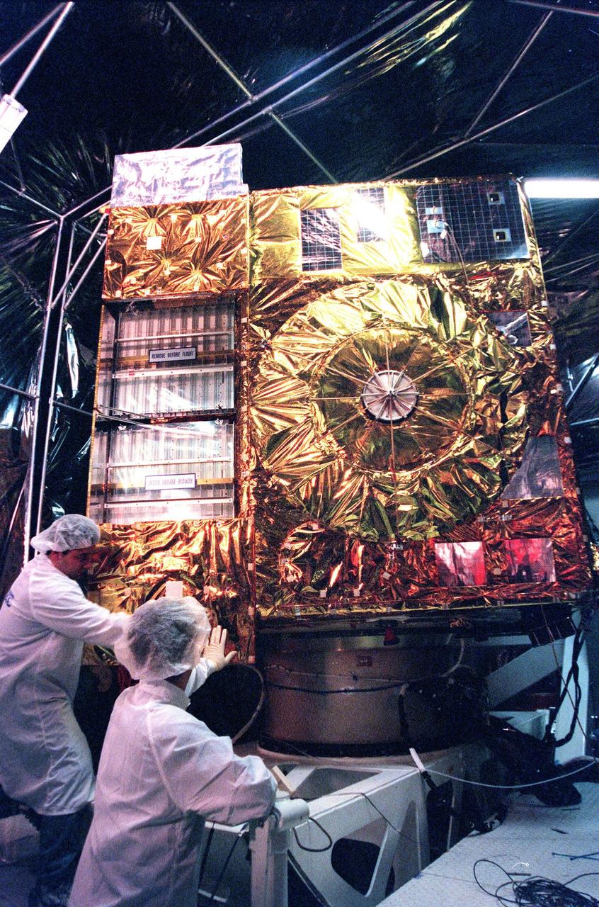 With the light casting a rosy glow in a specially built clean room at Astrotech, Titusville, Fla., Loral technicians Roberto Caballero (left) and Paul Giordano (right) maneuver the <a href="http://www-pao.ksc.nasa.gov/kscpao/captions/subjects/goes-l.htm">GOES-L</a> weather satellite into position for testing the deployment of the sounder instrument's cooler cover door. The sounder, one of two meteorological instruments on the satellite, measures temperature and moisture in a vertical column of air from the satellite to Earth. Its findings will help forecast weather. GOES-L, which is to be launched from Cape Canaveral Air Station aboard an Atlas II rocket in late March, is the fourth of a new advanced series of geostationary weather satellites for the National Oceanic and Atmospheric Administration. It is a three-axis inertially stabilized spacecraft that will provide pictures as well as perform the atmospheric sounding. Once launched, the satellite, to be designated GOES-11, will undergo checkout and provide backup capabilities for the existing, aging GOES East weather satellite