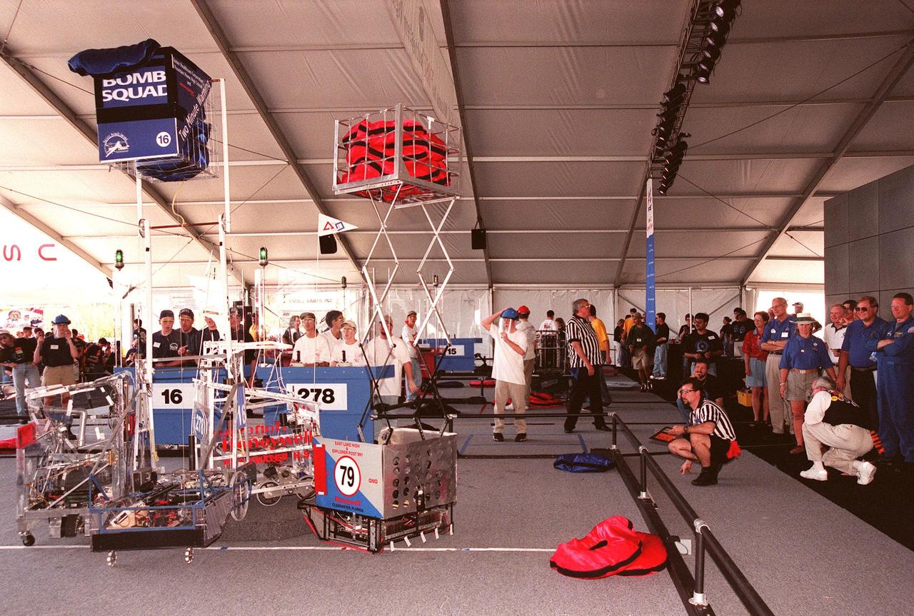 Four robots vie for position on the playing field during the 1999 FIRST Southeastern Regional robotic competition held at KSC. Powered by 12-volt batteries and operated by remote control, the robotic gladiators spent two minutes each trying to grab, claw and hoist large, satin pillows onto their machines. Student teams, shown behind protective walls, play defense by taking away competitors' pillows and generally harassing opposing machines. Two of the robots have lifted their caches of pillows above the field, a movement which earns them points. Along with the volunteer referees, at the edge of the playing field, judges at right watch the action. FIRST is a nonprofit organization, For Inspiration and Recognition of Science and Technology. The competition comprised 27 teams, pairing high school students with engineer mentors and corporations. The FIRST robotics competition is designed to provide students with a hands-on, inside look at engineering and other professional careers