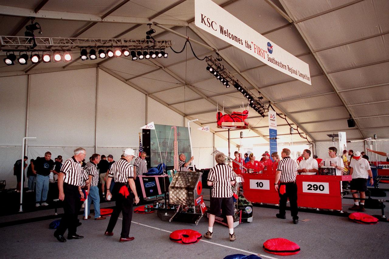 Referees check the robots on the floor of the playing field after a qualifying match of the 1999 Southeastern Regional robotic competition at Kennedy Space Center Visitor Complex . Thirty schools from around the country have converged at KSC for the event that pits gladiator robots against each other in an athletic-style competition. The robots have to retrieve pillow-like disks from the floor, as well as climb onto the platform (with flags) and raise the cache of pillows to a height of eight feet. KSC is hosting the event being sponsored by the nonprofit organization For Inspiration and Recognition of Science and Technology, known as FIRST. The FIRST robotics competition is designed to provide students with a hands-on, inside look at engineering and other professional careers
