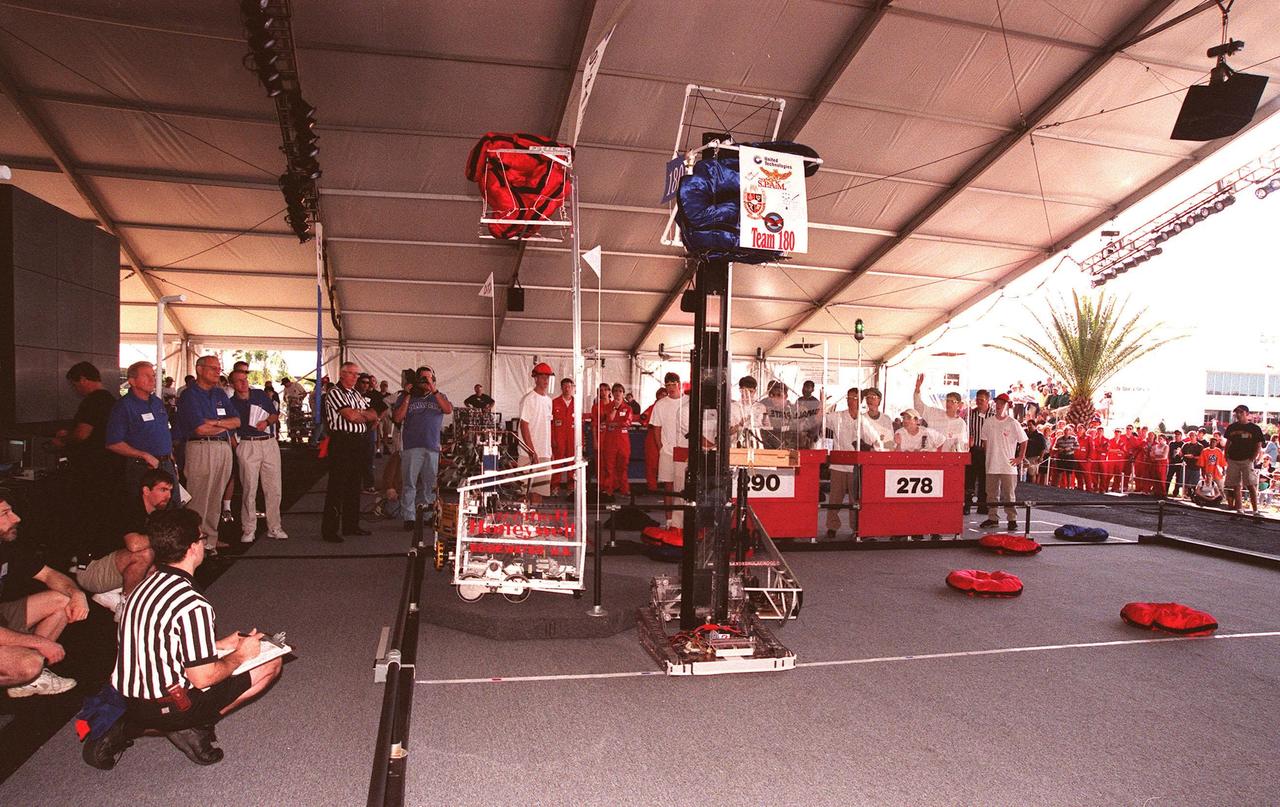During final matches at the 1999 Southeastern Regional robotic competition at the KSC Visitor Complex, referees and judges (blue shirts at left) watch as two robots raise their pillow disks to a height of eight feet, one of the goals of the competition. KSC Deputy Director for Launch and Payload Processing Loren Shriver is one of the judges. Thirty schools from around the country have converged at KSC for the event that pits gladiator robots against each other in an athletic-style competition. The robots have to retrieve the disks from the floor, climb onto a platform (with flags), as well as raise the cache of pillows, maneuvered by student teams behind protective walls. KSC is hosting the event being sponsored by the nonprofit organization For Inspiration and Recognition of Science and Technology, known as FIRST. The FIRST robotics competition is designed to provide students with a hands-on, inside look at engineering and other professional careers by pairing engineers and corporations with student teams