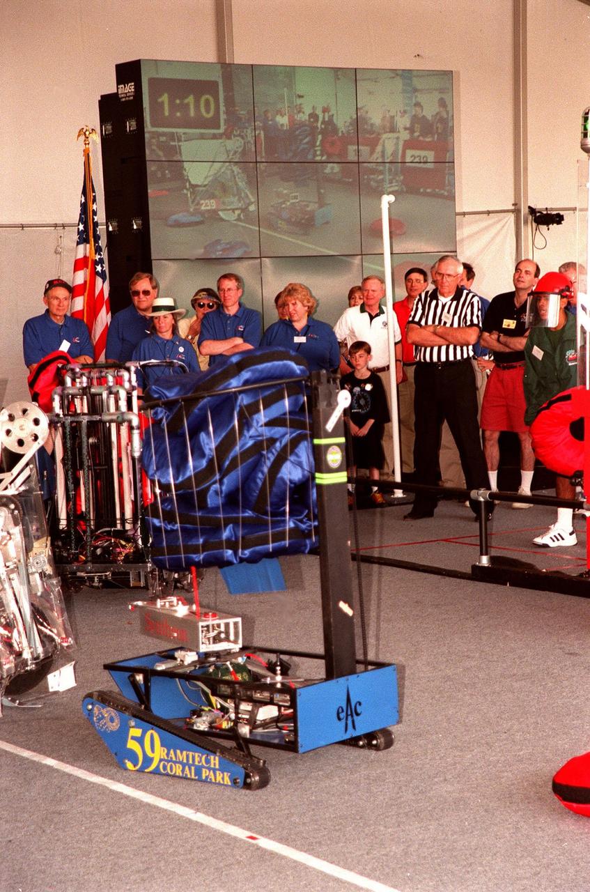 During the 1999 FIRST Southeastern Regional robotic competition held at KSC, a robot carrying its cache of pillow-like disks maneuvers to move around another at left. Powered by 12-volt batteries and operated by remote control, the robotic gladiators spend two minutes each trying to grab, claw and hoist the pillows onto their machines. Teams play defense by taking away competitors' pillows and generally harassing opposing machines. Behind the field are a group of judges, including KSC former KSC Director of Shuttle Processing Robert Sieck (left, in cap), and Center Director Roy Bridges (in white shirt). A giant screen TV in the background displays the action on the playing field. FIRST is a nonprofit organization, For Inspiration and Recognition of Science and Technology. The competition comprised 27 teams, pairing high school students with engineer mentors and corporations. The FIRST robotics competition is designed to provide students with a hands-on, inside look at engineering and other professional careers