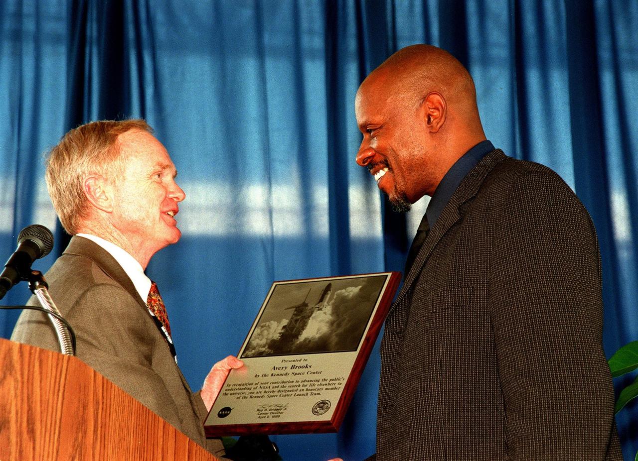 KENNEDY SPACE CENTER, FLA. -- At the grand opening of the newly expanded KSC Visitor Complex, Center Director Roy Bridges presents Deep Space Nine star Avery Brooks with a plaque, recognizing his contribution to advancing the public's understanding of NASA and the search for life elsewhere in the universe. Brooks narrates the new film Quest for Life at the Visitor Center. The $ 13 million addition to the Visitor Complex now includes an International Space Station-themed ticket plaza, featuring a structure of overhanging solar panels and astronauts performing assembly tasks, a new information center, films, and exhibits. The KSC Visitor Complex was inaugurated three decades ago and is now one of the top five tourist attractions in Florida. It is located on S.R. 407, east of I-95, within the Merritt Island National Wildlife Refuge
