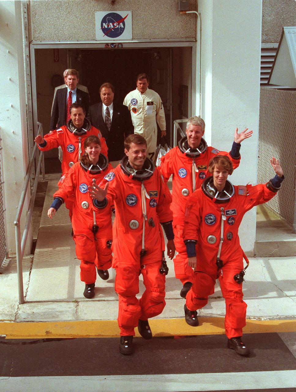 After suiting up for their practice countdown exercise, STS-93 crew members wave to onlookers as they head for the bus outside the Operations and Checkout Building for the trip to Launch Pad 39B. From rear left are Mission Specialists Michel Tognini of France, who represents the Centre National d'Etudes Spatiales (CNES), and Catherine G. Coleman (Ph.D.), Pilot Jeffrey S. Ashby, Mission Specialist Steven A. Hawley (Ph.D.), and Commander Eileen M. Collins. Collins is the first woman to serve as a Shuttle commander. The crew members have been taking part in a Terminal Countdown Demonstration Test, which familiarizes them with the mission, provides training in emergency exit from the orbiter and launch pad, and includes a launch-day dress rehearsal culminating with a simulated main engine cut-off. The primary mission of STS-93 is the release of the Chandra X-ray Observatory, which will allow scientists from around the world to obtain unprecedented X-ray images of exotic environments in space to help understand the structure and evolution of the universe. The targeted launch date for STS-93 is no earlier than July 20 at 12:36 a.m. EDT from Launch Pad 39B